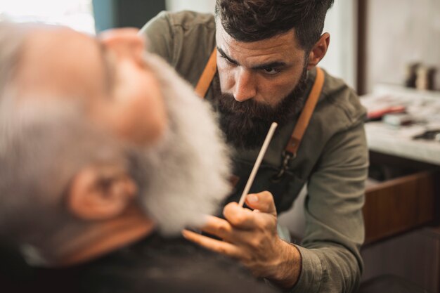 Parrucchiere maschio dei pantaloni a vita bassa che lavora con la barba del cliente senior