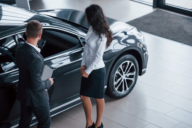 Parte anteriore dell'auto. Cliente femminile e uomo d'affari barbuto elegante moderno nel salone dell'automobile