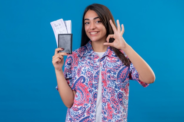 Passaporto turistico della holding della giovane bella donna con i biglietti che guarda l'obbiettivo sorridente allegramente facendo segno giusto in piedi sopra fondo blu isolato