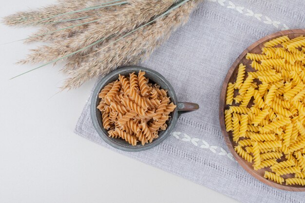 Pasta a spirale cruda in tazza e piatto sulla tovaglia con grano.