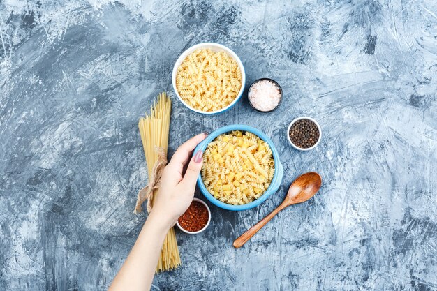 Pasta assortita in ciotole e mano che tiene una ciotola di pasta con spezie, vista dall'alto cucchiaio di legno su uno sfondo di gesso grigio