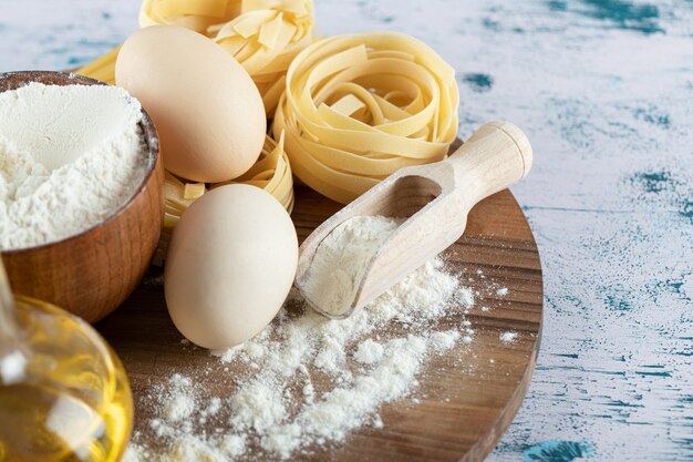 Pasta cruda con olio, uovo e ciotola di farina su tavola di legno.