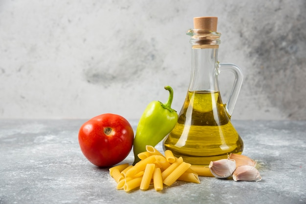 Pasta cruda di penne con bottiglia di olio d'oliva e verdure sulla tavola di marmo. Avvicinamento.