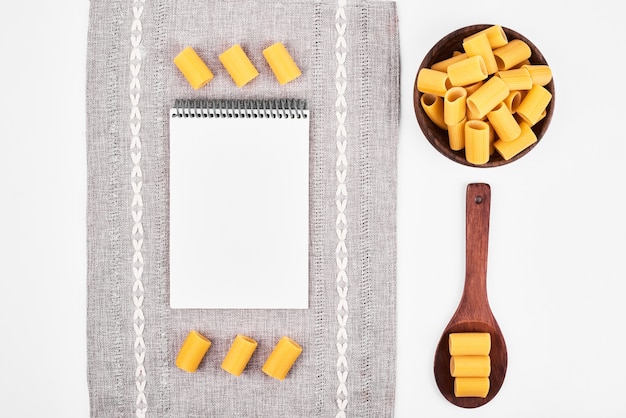 Pasta cruda in cucchiai di legno su sfondo bianco con un ricettario.