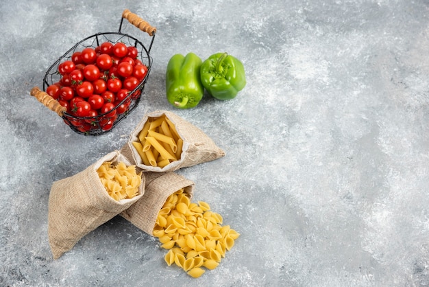 Pasta cruda in un sacchetto rustico con pomodorini e peperoni su tavola di marmo.