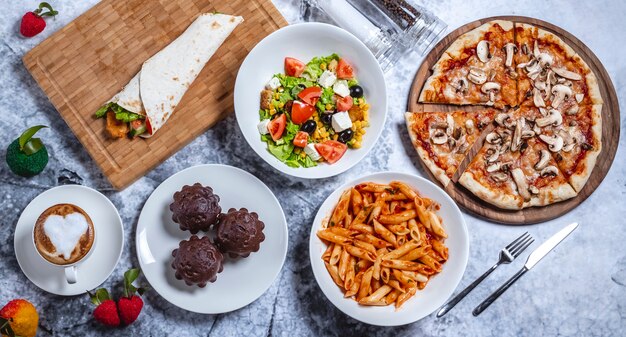 Pasta e tazza di caffè del penne dei muffin del cioccolato del rotolo di pollo della pizza del fungo dell'insalata greca degli alimenti a rapida preparazione di vista superiore sul tavolo