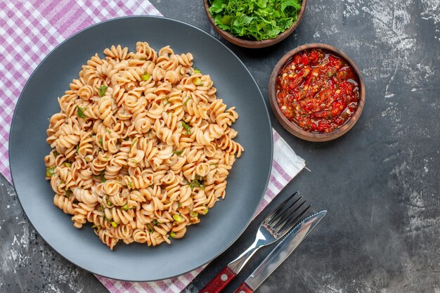 Pasta rotini vista dall'alto sulla piastra sul tovagliolo a scacchi bianco rosa forchetta e coltello verdure tritate e salsa di pomodoro in ciotole sul tavolo grigio