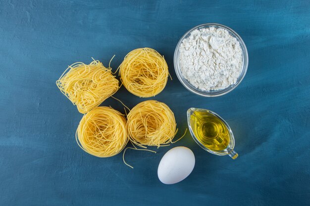 Pasta secca cruda del nido con uovo crudo e olio su una superficie blu scuro.