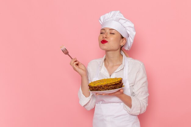 Pasticcere femminile di vista frontale in abbigliamento bianco che tiene i pasticcini deliziosi che lo assaggiano sulla scrivania rosa