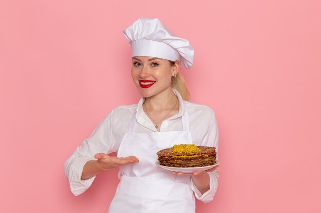 Pasticcere femminile di vista frontale in usura bianca che tiene i pasticcini deliziosi sul lavoro di lavoro della pasticceria dolce della pasticceria della parete rosa chiaro