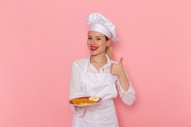 Pasticcere femminile di vista frontale nel piatto della tenuta di usura bianca con il cibo sul cibo della cucina della cucina di lavoro del cuoco della parete rosa chiaro