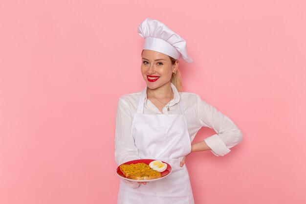 Pasticcere femminile di vista frontale nel piatto della tenuta di usura bianca con il cibo sul cibo della cucina della cucina di lavoro del cuoco della parete rosa