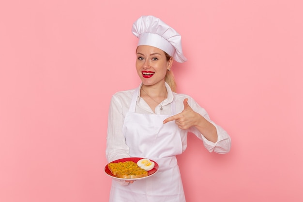 Pasticcere femminile di vista frontale nel piatto della tenuta di usura bianca con il cibo sul cibo di cucina della cucina di lavoro della parete rosa