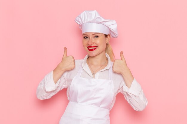 Pasticcere femminile di vista frontale nell'usura bianca che sorride e che posa sul lavoro rosa della pasticceria della pasticceria del cuoco della parete