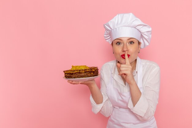 Pasticcere femminile di vista frontale nell'usura bianca che tiene i pasticcini deliziosi che posano sulla pasticceria dolce della pasticceria della parete rosa