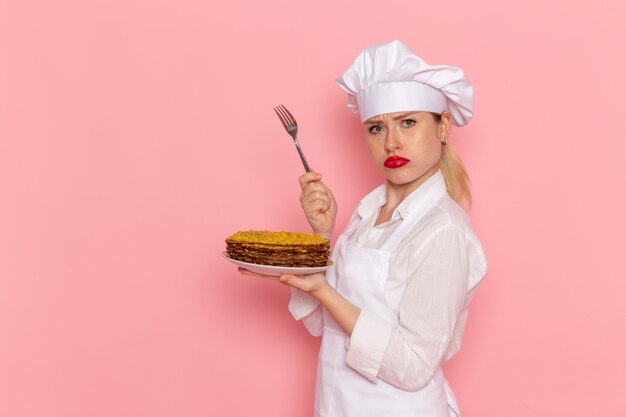 Pasticcere femminile di vista frontale nell'usura bianca che tiene i pasticcini e la forcella deliziosi sulla pasticceria rosa della parete che cucina la pasticceria della torta