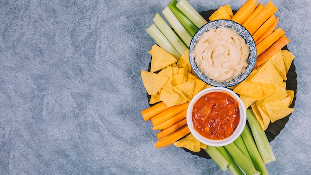 Patatine al nachos messicano; fette di carota con gambo di sedano in vassoio con salsa di salsa in ciotola su sfondo di cemento