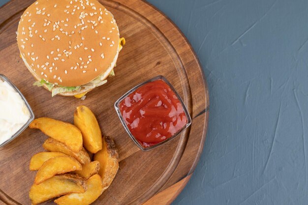 Patatine fritte con ciotole di ketchup e mayonaisse, accanto a un hamburger su tavola di legno su sfondo blu. Foto di alta qualità