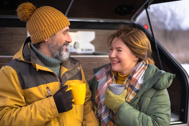 Pausa caffè durante il viaggio coppia matura momenti di relax insieme al mini camper Coppia di mezza età bere dalle tazze nido in piedi a un camper Viaggio avventura in famiglia Tempo divertente Concetto di viaggio in famiglia