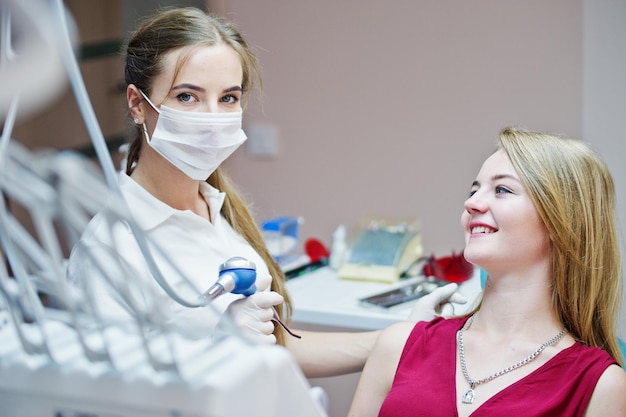 Paziente attraente in abito rosso viola sdraiato sulla poltrona del dentista mentre il dentista femminile si cura i denti con strumenti speciali