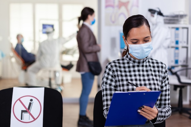 Paziente di stomatologia nell'area di attesa del denstiry che compila il modulo prima della consultazione con il dentista vestito con tuta in dpi come precauzione di sicurezza contro l'infezione da coronavirus durante l'epidemia globale.