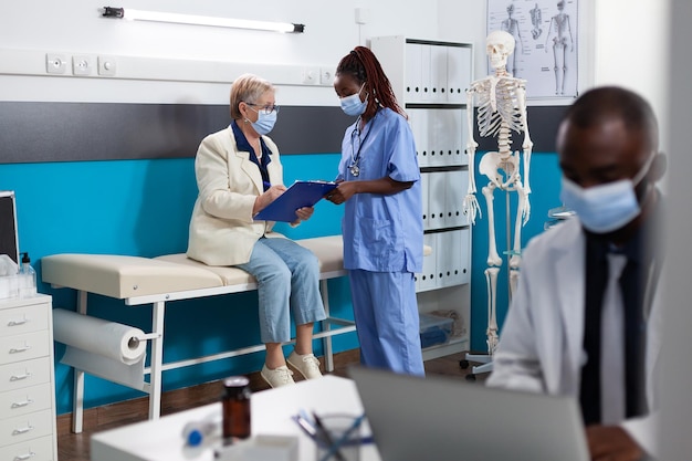 Paziente in pensione con maschera facciale di protezione che firma documenti medici durante l'appuntamento medico nell'ufficio dell'ospedale. Infermiera terapista che spiega la diagnosi di malattia discutendo il trattamento farmacologico