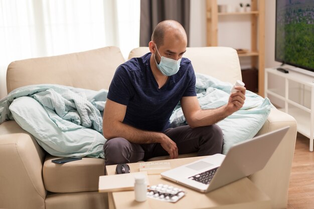 Paziente maschio che tiene la bottiglia di pillole guardando il laptop durante l'autoisolamento.