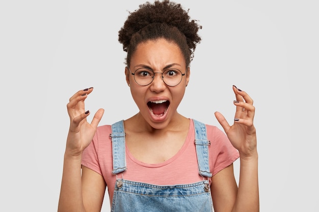 Pazza e furiosa giovane donna dalla pelle scura gesticola con rabbia, urla di stress, indossa abiti casual, si erge contro il muro bianco. La femmina afroamericana esprime rabbia, ha un'espressione malvagia.