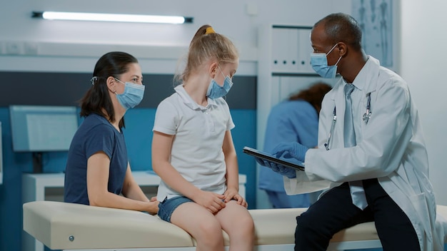 Pediatra utilizzando tablet per prendere appunti all'appuntamento di consultazione in ufficio. Medico maschio che tiene il dispositivo e discute con il bambino e la madre, fornendo assistenza medica.