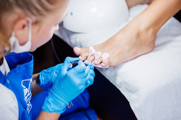 Pedicure da salone domestico Trattamento per la cura dei piedi e unghie Il processo di pedicure professionale Il maestro in guanti blu applica lo smalto gel rosa chiaro