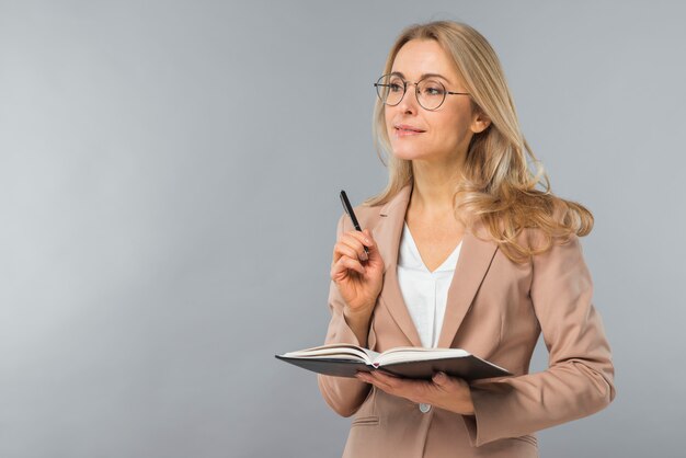 Penna e diario biondi sorridenti sicuri della tenuta della giovane donna a disposizione contro fondo grigio