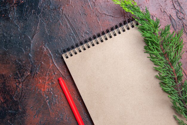 Penna rossa vista dall'alto un blocco note con piccolo fiocco un ramo di un albero di pino su superficie rosso scuro con spazio libero