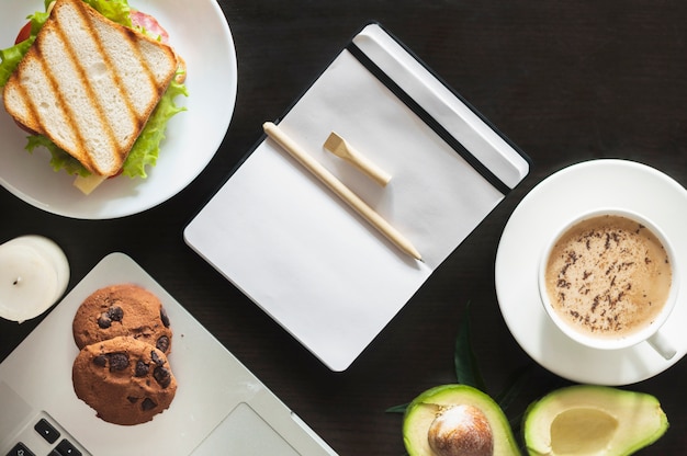 Penna su diario vuoto con sandwich; biscotti; tazza di caffè e avocado su sfondo nero