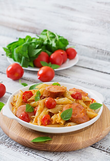 Penne al sugo di pomodoro con salsiccia, pomodori, basilico verde decorato in padella su un tavolo di legno