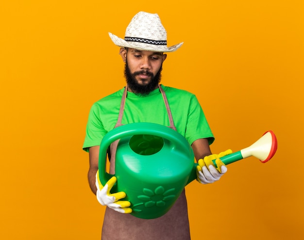 Pensando a un giovane giardiniere afroamericano che indossa cappello e guanti da giardinaggio che tengono e guardano l'annaffiatoio