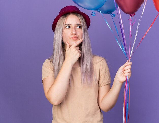 Pensando a una giovane bella ragazza che indossa un cappello da festa che tiene palloncini e ha afferrato il mento