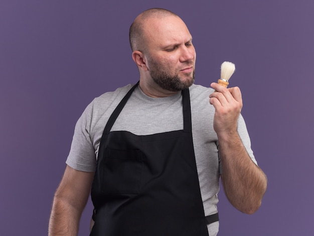 Pensando barbiere maschio di mezza età in tenuta uniforme e guardando il pennello da barba isolato su parete viola