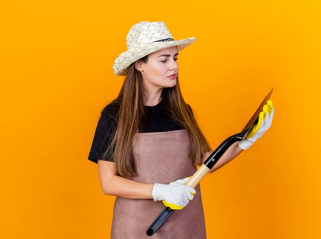 Pensando bella ragazza giardiniere che indossa uniforme e cappello da giardinaggio con guanti che tengono e guardando vanga isolato su sfondo arancione