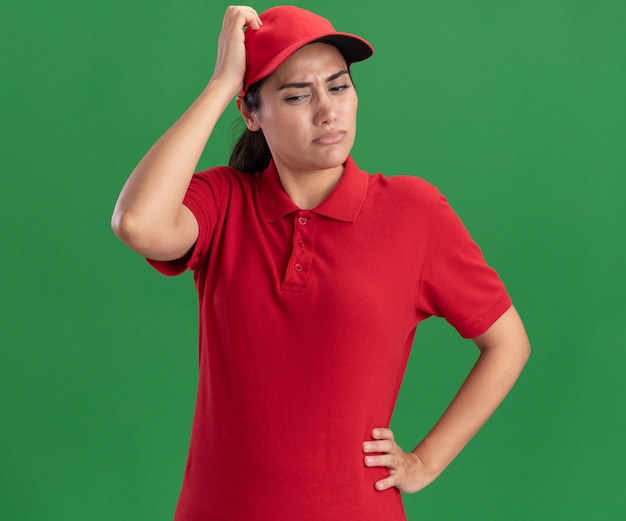 Pensando che guarda giù la giovane ragazza di consegna che indossa l'uniforme e il cappuccio che graffia la testa isolata sulla parete verde