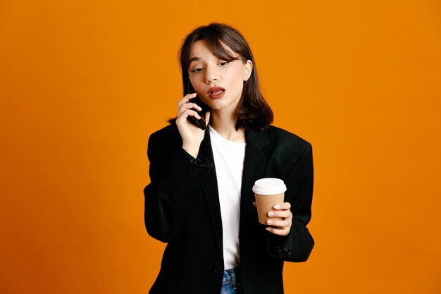 Pensando che tiene una tazza di caffè parla al telefono giovane bella donna che indossa una giacca nera isolata su sfondo arancione