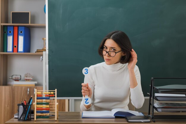 pensando di mettere la mano sulla testa giovane insegnante femminile che indossa occhiali tenendo il numero fan seduto alla scrivania con gli strumenti della scuola in classe