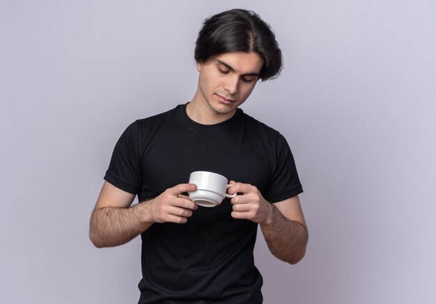 Pensando giovane bel ragazzo che indossa la maglietta nera che tiene e guardando la tazza di caffè isolato sul muro bianco