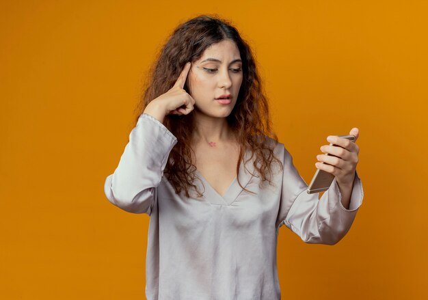 Pensando giovane bella ragazza che tiene e guardando il telefono e mettendo il dito sulla testa isolata sulla parete gialla