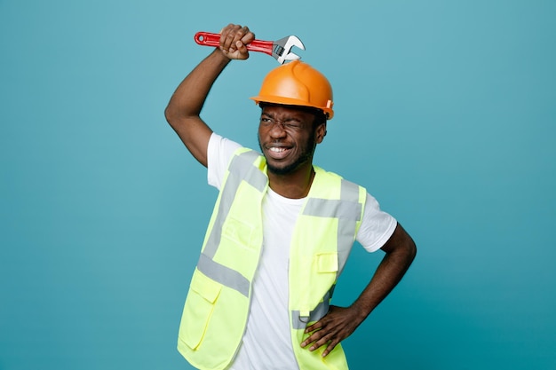 Pensando giovane costruttore afroamericano in uniforme tenendo la chiave a gas sulla testa isolata su sfondo blu