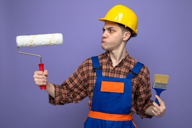 Pensando giovane costruttore maschio indossando uniforme tenendo il pennello guardando la spazzola a rullo in mano