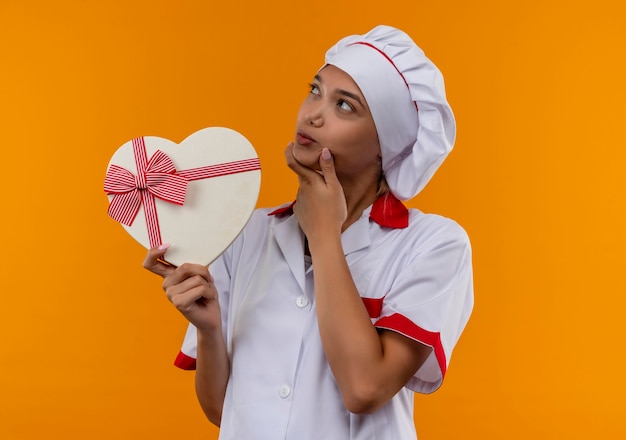 Pensando giovane cuoco femmina che indossa lo chef uniforme tenendo la scatola a forma di cuore mettendo la mano sul mento isolato su sfondo arancione