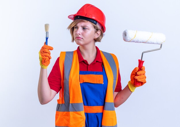 Pensando giovane donna costruttore in uniforme tenendo la spazzola a rullo e guardando il pennello in mano isolata sul muro bianco