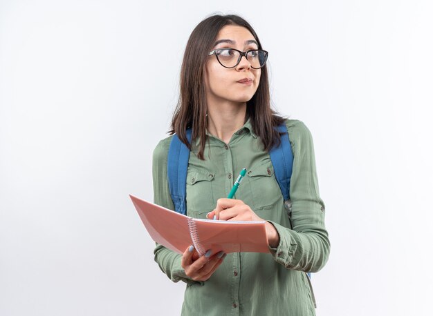 Pensando giovane donna della scuola con gli occhiali con lo zaino che scrive qualcosa sul taccuino