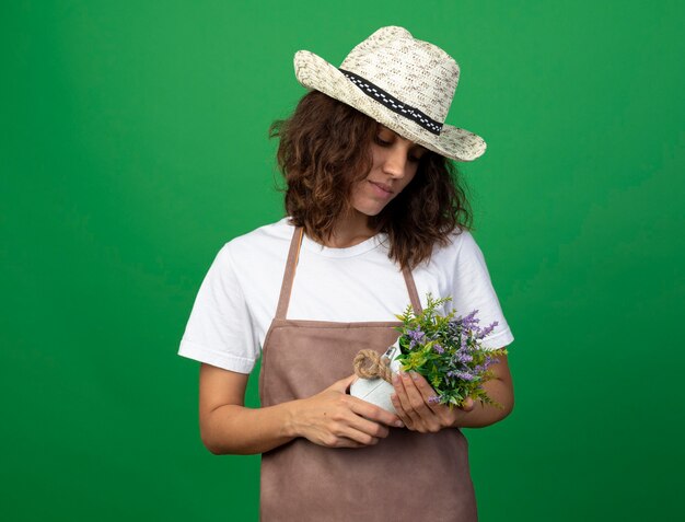 Pensando giovane giardiniere femminile in uniforme che indossa cappello da giardinaggio tenendo e guardando il fiore in vaso di fiori