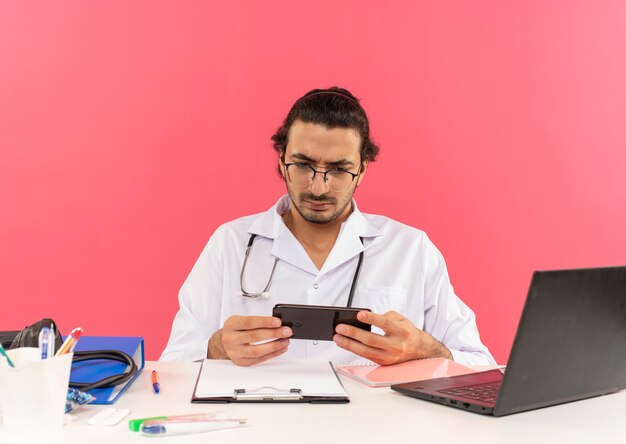 Pensando giovane medico maschio con occhiali medici che indossano accappatoio medico con stetoscopio seduto alla scrivania